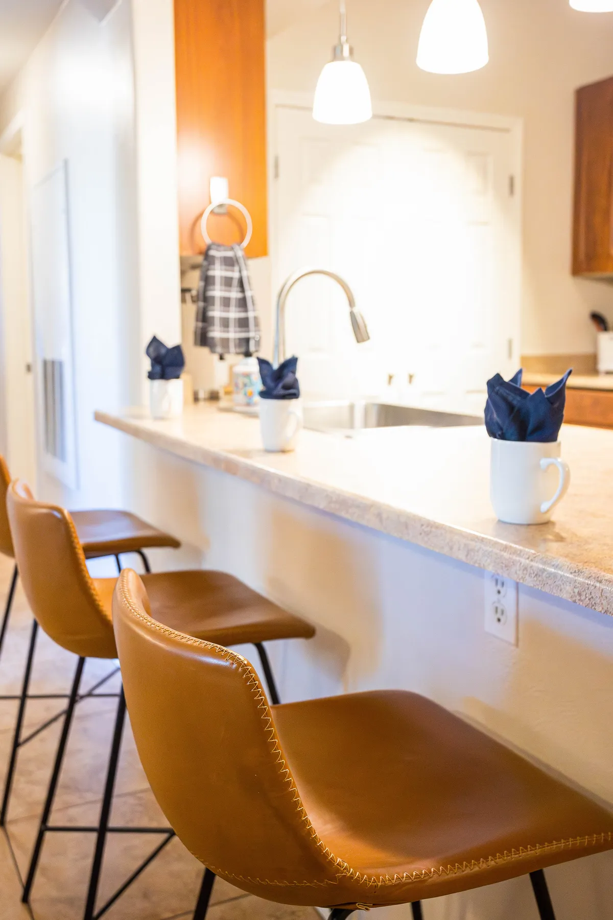 A warm, welcoming kitchen scene featuring brown leather bar stools at a granite countertop, set with white mugs and navy napkins. Friends share a laugh, highlighting the casual, community-driven atmosphere ideal for socializing or enjoying a morning coffee.