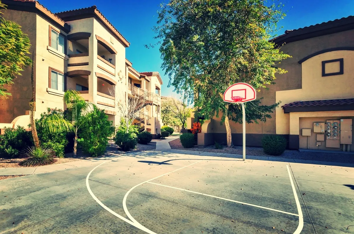 Exterior community area with a basketball hoop, lush greenery, and apartment buildings. The scene is lively and reflects communal outdoor spaces where residents can socialize, unwind, or include their pets in the fun.