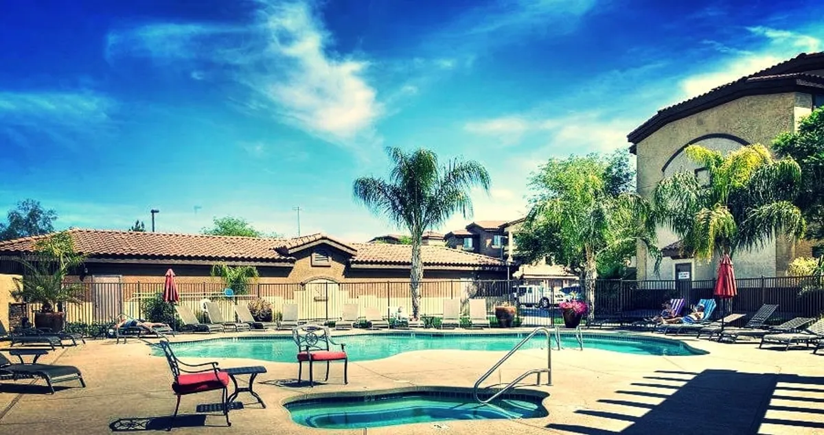 A sparkling swimming pool flanked by lounge chairs and palm trees, with pops of color from red umbrellas. The scene is bright and inviting, capturing a quintessential Arizona resort-style leisure area. People can envision themselves swimming, sunbathing, or socializing poolside under sunny skies.