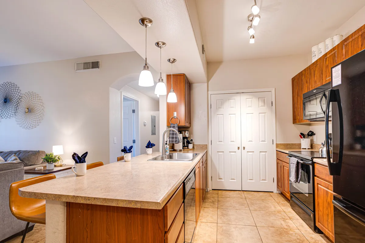 A modern kitchen island is the focal point, surrounded by stainless steel appliances, pendant lighting, and decorative wall art. The spacious design encourages cooking, entertaining, and gathering with friends and neighbors.