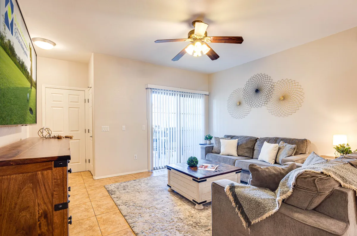 A cozy living area features a sofa, coffee table, decorative wall art, and a sliding glass door that brings in abundant sunlight. The door leads out to the private balcony, and garden views are visible through the wide windows, reinforcing the connection between indoor comfort and the inviting outdoor space.