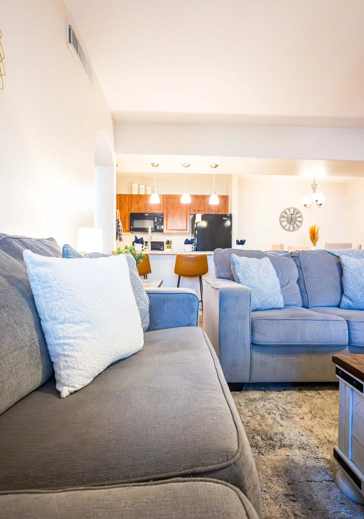This image showcases a spacious open-concept living area with comfortable sofas, a dining area with a table and chairs, the kitchen in full view, decorative plants, and a wall clock. Natural light streams in, creating a welcoming atmosphere ideal for gatherings and relaxation.