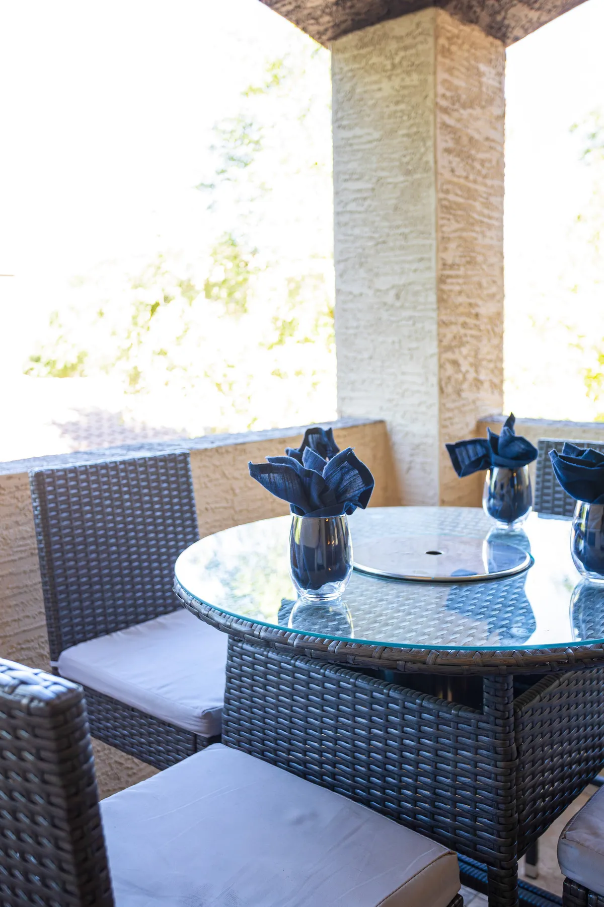 An outdoor dining setup featuring a glass table with woven chairs and decorative napkins, all bathed in natural light. The ambiance suggests a space perfect for BBQs, al fresco meals, or evening gatherings with friends and neighbors.