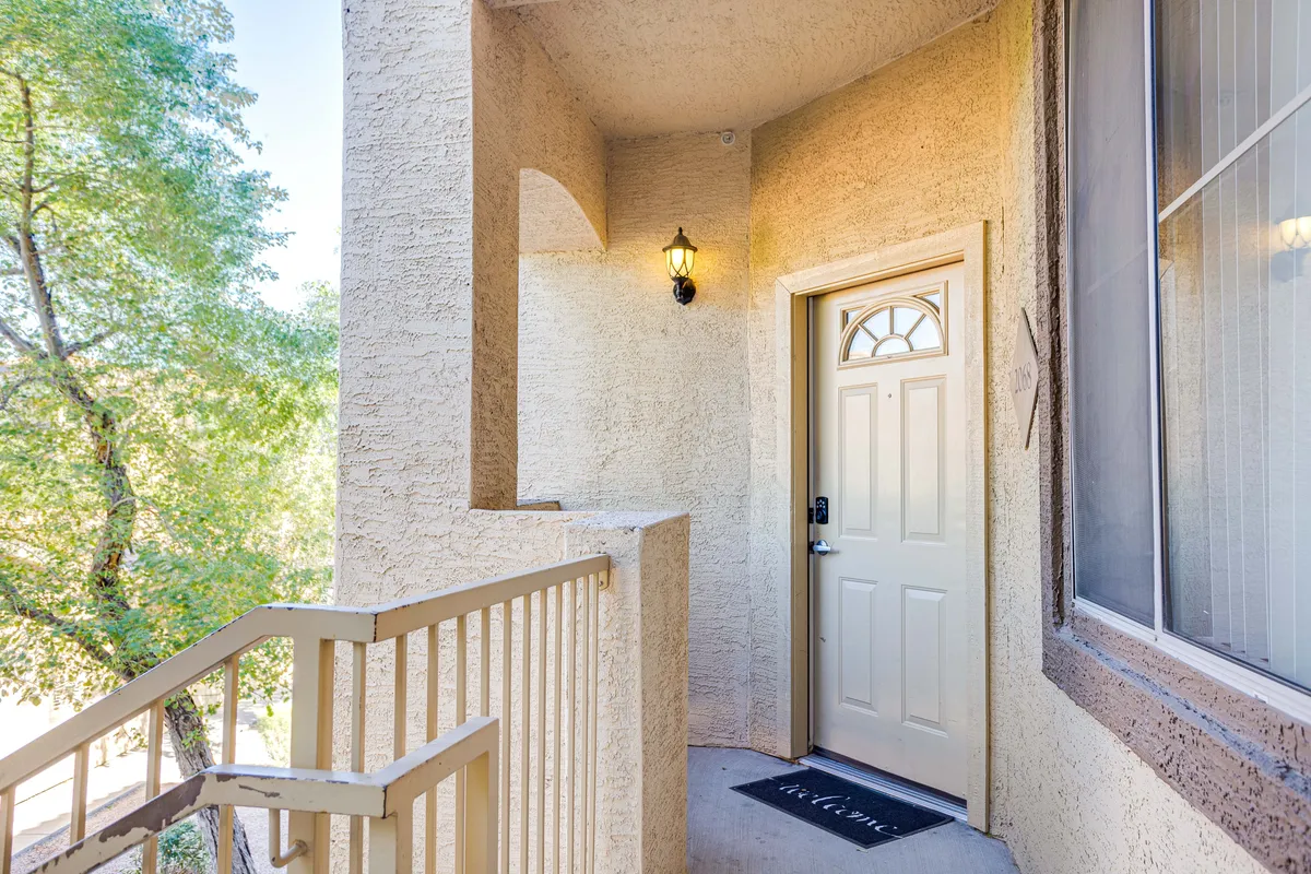 This exterior image highlights the inviting entryway of the property, featuring a welcoming front door, stylish light fixture, stair railing, and lush surrounding greenery. The porch area suggests a transition space between outdoor activities and indoor comforts, reinforcing the connection to the community and amenities nearby.