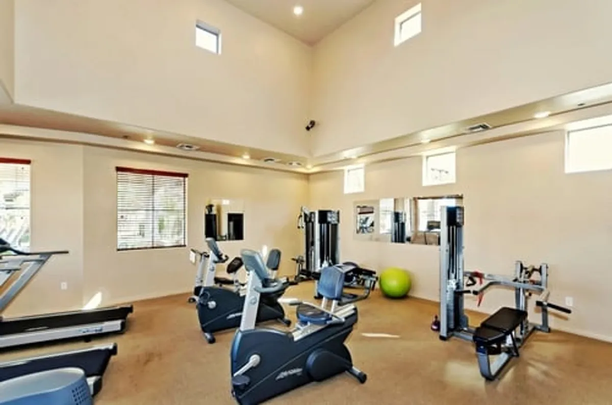 The image shows a well-equipped fitness room with modern exercise equipment, large mirrors for form checks, big windows letting in natural light, and a green exercise ball. The space feels energized and motivating, perfect for guests with an active lifestyle.