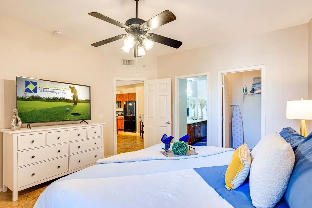 A cozy bedroom featuring a well-dressed bed with decorative pillows, a dresser adorned with decorations, and a television displaying a tranquil scene. The room feels comfortable and suitable for relaxing or unwinding with a show.
